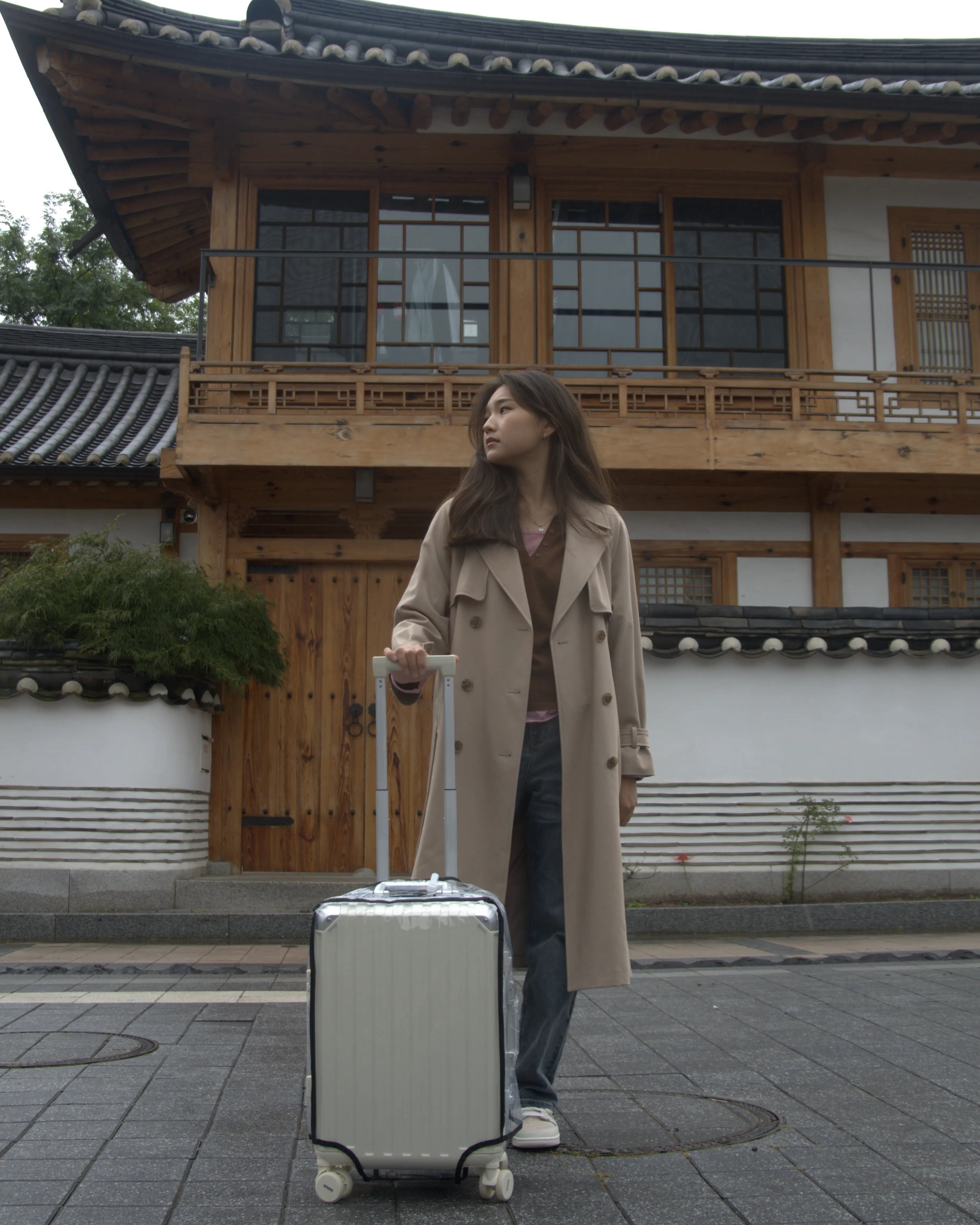 Woman in a trench coat standing next to a traditional building with a suitcase.