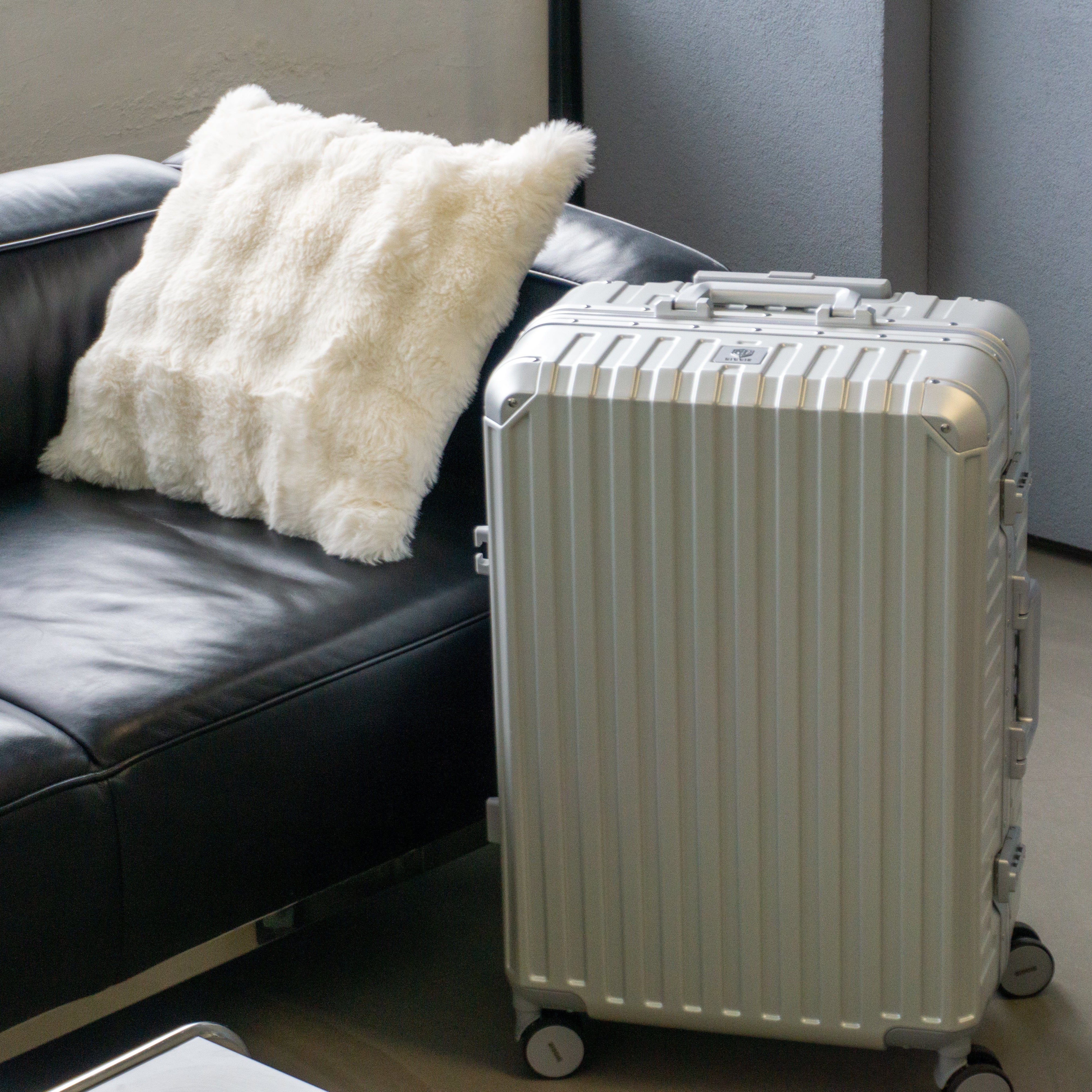 Titanium Silver luggage on a floor next to a black leather couch with a white textured pillow.